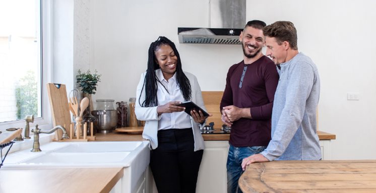 Smiling couple in modern kitchen discussing mortgage options with a friendly home loan advisor using a digital tablet.