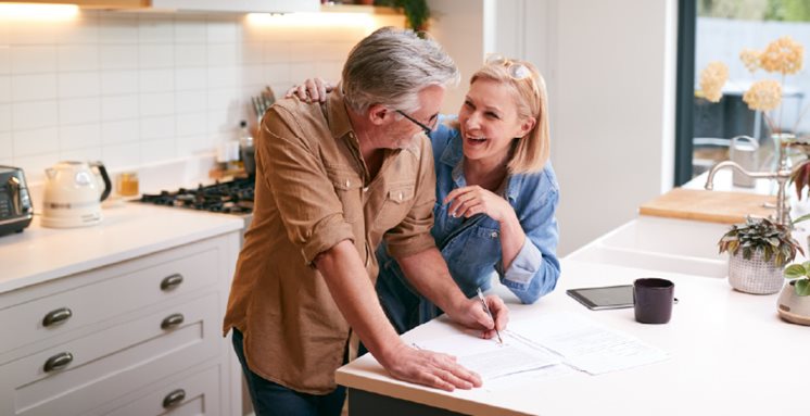 A happy middle-aged couple reviews and signs home refinancing paperwork together in a bright modern kitchen.