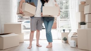 First-time homebuyers in Kansas. Two people holding boxes in a bright room filled with moving boxes and potted plants, suggesting a home move-in scene.
