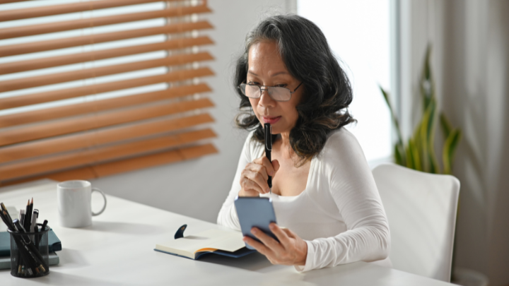 Woman seated at a desk, looking at her smartphone with a notebook nearby